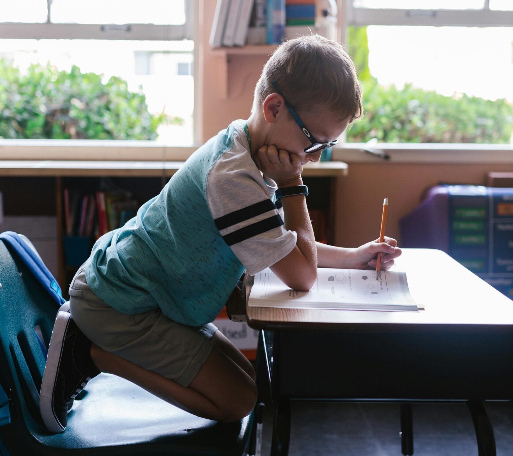 child sitting on knees in chair at school desk working on school work